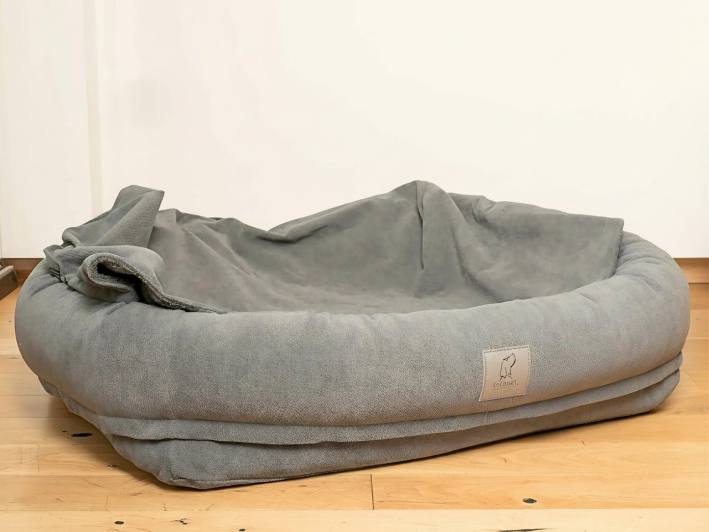 Grey bolster dog bed with hood folded down on wooden floor.