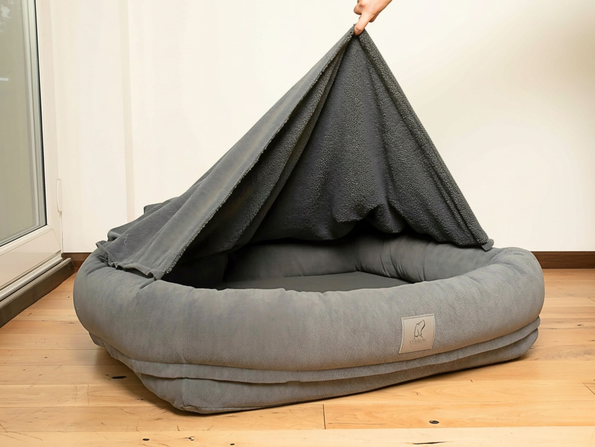 Grey bolster dog bed with removable zip-on hood lifted by hand.