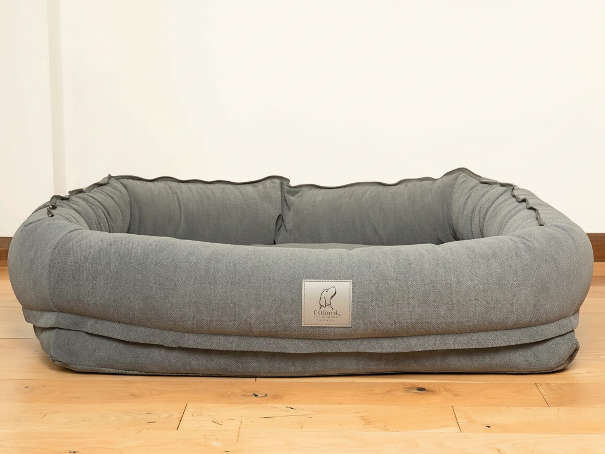 Grey bolster dog bed without hood, shown empty on wooden floor.