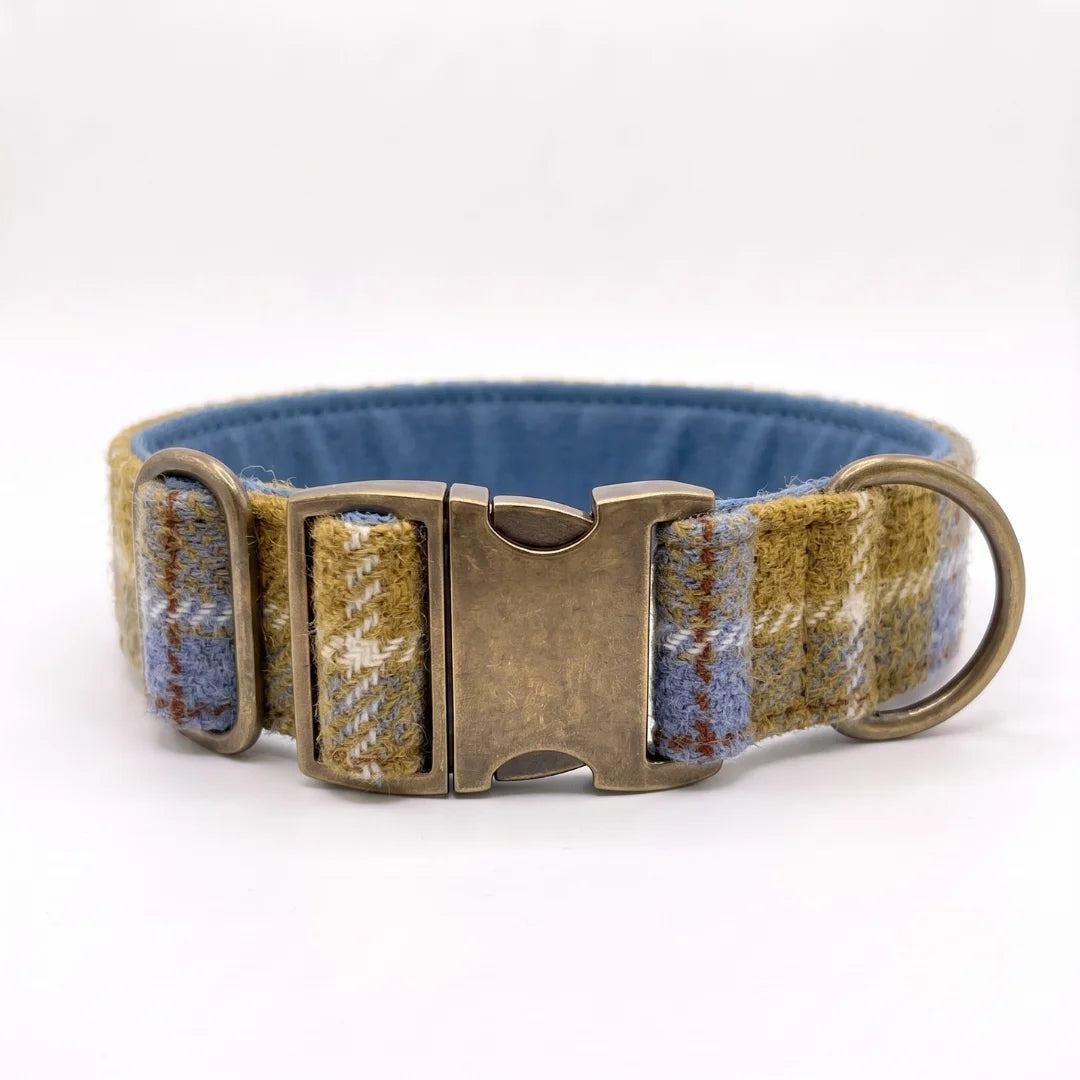 A woven dog collar with a blue interior, featuring a mustard and blue plaid pattern on the exterior, a bronze-toned metal buckle, and a matching metal ring.