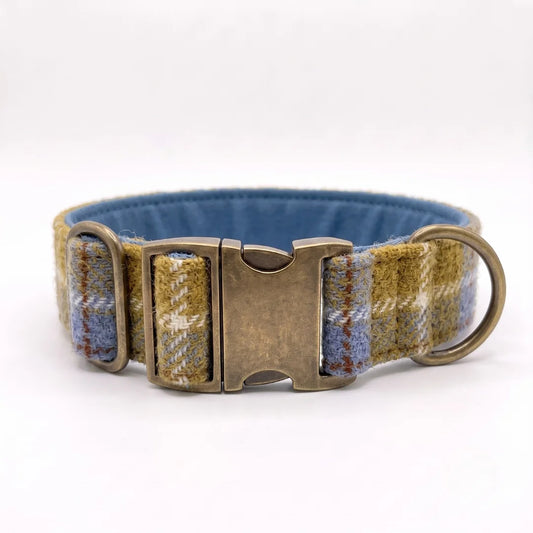 A woven dog collar with a blue interior, featuring a mustard and blue plaid pattern on the exterior, a bronze-toned metal buckle, and a matching metal ring.
