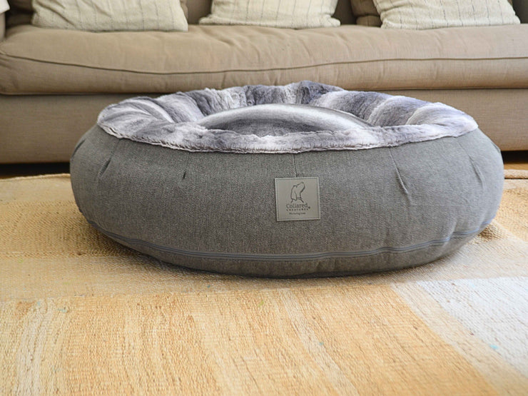 Grey calming donut dog bed with soft faux fur lining, placed in a cosy living room setting with a beige sofa in the background. Branded label from Collared Creatures visible on the front.