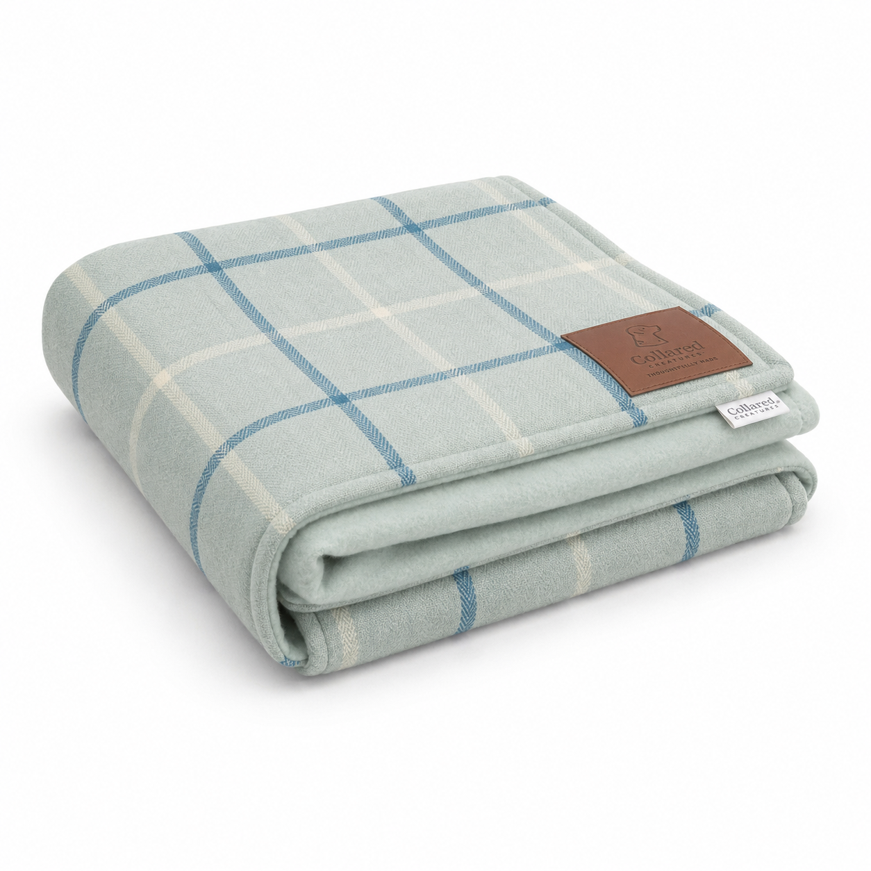 A folded plaid blanket in a light sage green, cream, and blue colorway, featuring a brown leather brand patch on the corner.