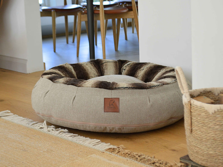 Beige donut dog bed with brown faux fur inner lining by Collared Creatures, placed on a wooden floor next to a woven basket in a modern home.