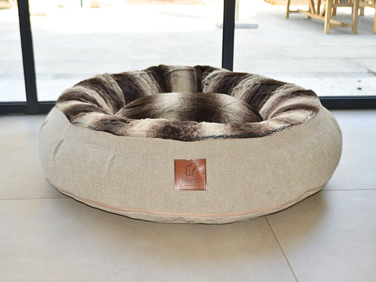 Large beige donut dog bed with deep brown faux fur inner lining by Collared Creatures, positioned in front of patio doors on a tiled floor.