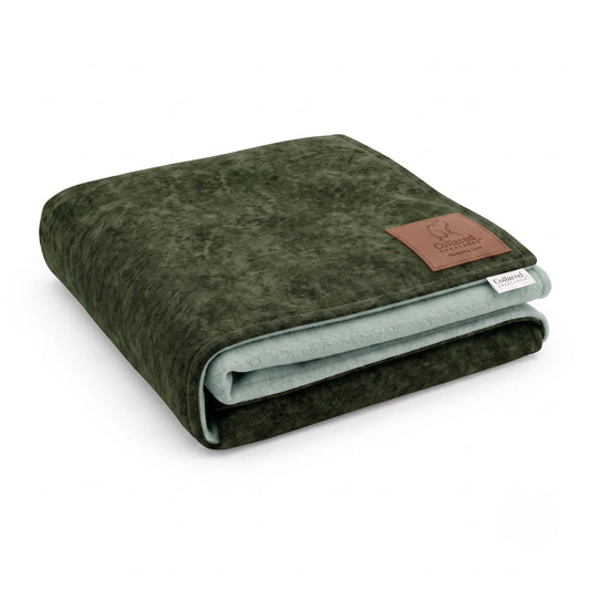 A folded plush blanket in a dark olive green color, showing a light sage green inner side and a brown leather brand patch on the corner.