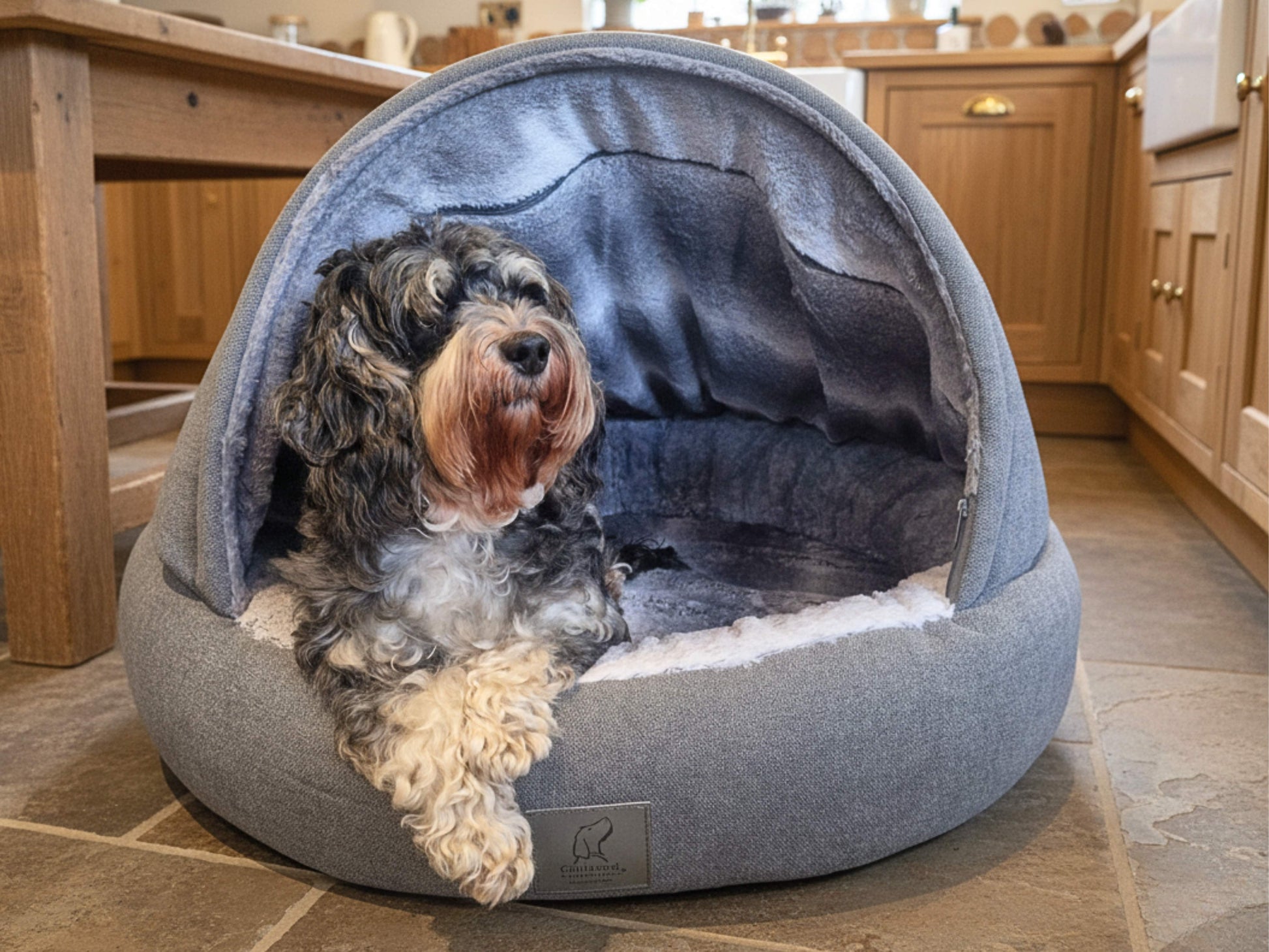 Dog relaxing in a grey Collared Creatures cave dog bed placed in a cozy country kitchen with wooden cabinets and stone floor
