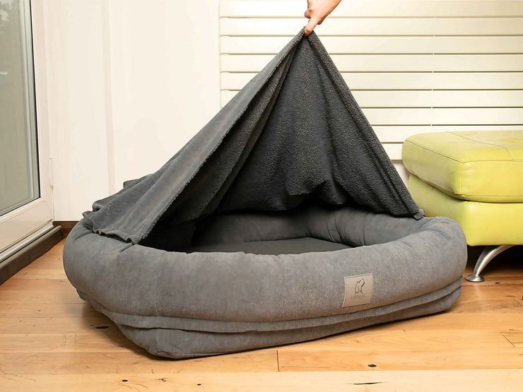 Grey bolster dog bed with removable hood partially lifted to show inner teddy fleece lining