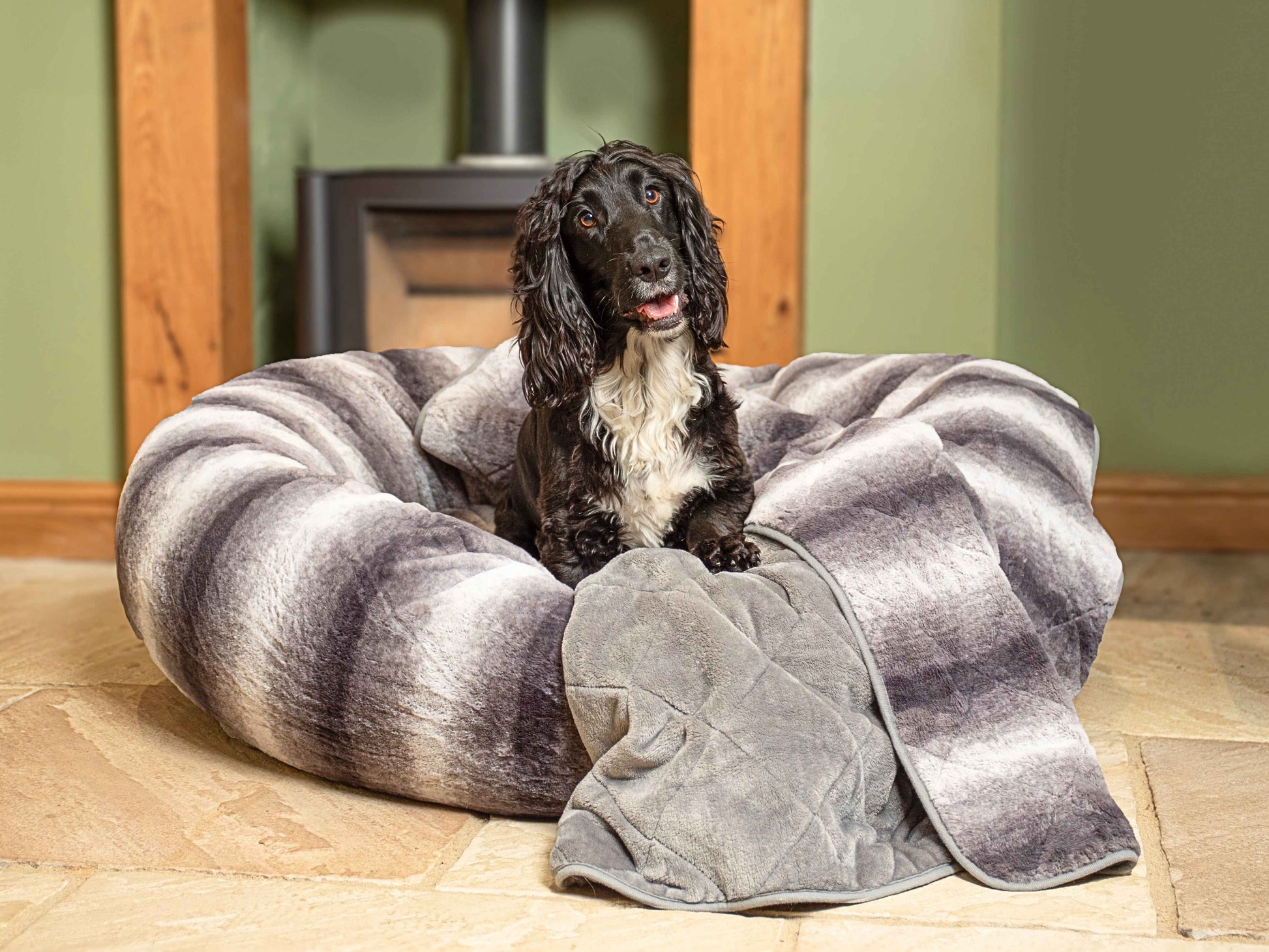 Happy dog resting on luxury grey dog blanket placed inside a plush dog bed by the fireplace.