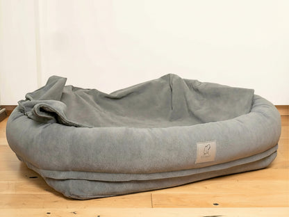 Grey bolster dog bed with hood folded down on wooden floor.