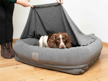 Spaniel resting inside a grey bolster dog bed while the removable hood is partially lifted.